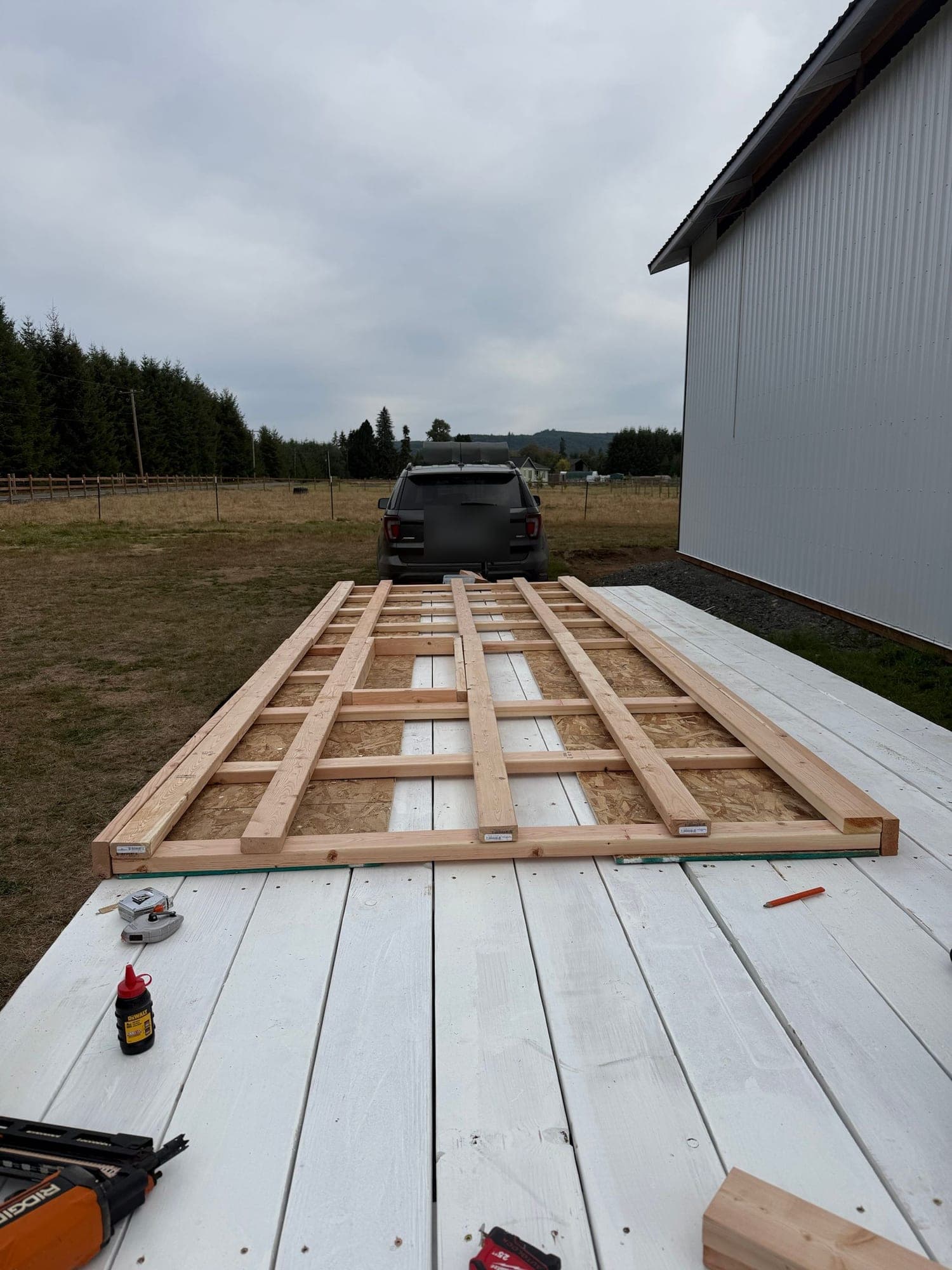 A wall frame of fresh lumber laid out on the white-painted trailer deck next to the farm's pole barn